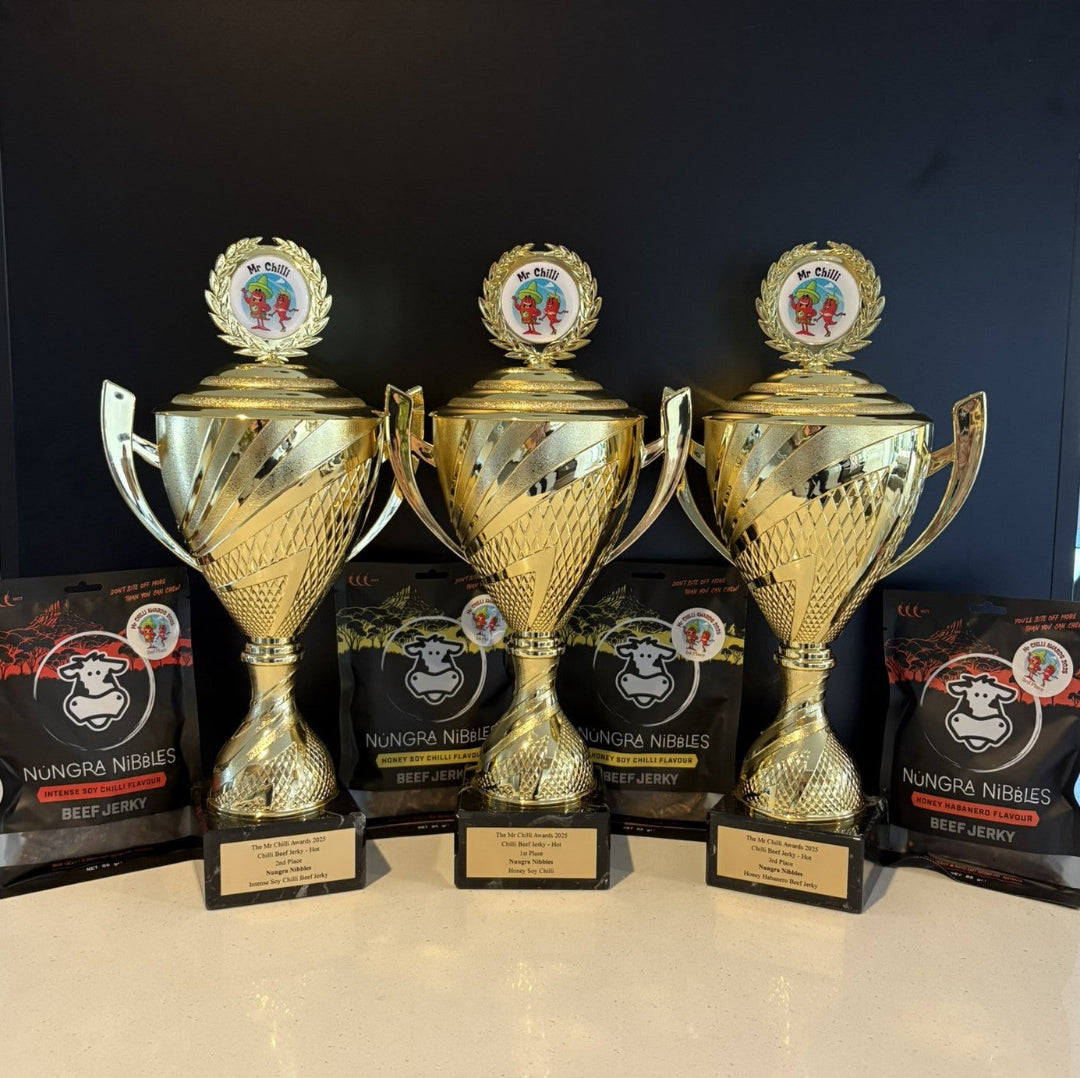 Three gold trophies on a black background with Nungra Nibbles beef jerky boxes.