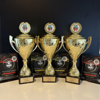 Three gold trophies on a black background with Nungra Nibbles beef jerky boxes.